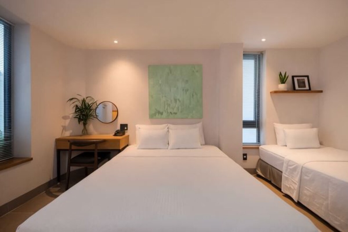 A bedroom with a large bed, a desk, a chair, a green abstract painting, and a window with blinds.