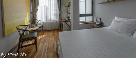 Une chambre d’hôtel avec un grand lit, une table à manger avec des chaises, un petit bureau avec une chaise, une fenêtre avec des rideaux et une étagère avec des photos et une plante.
