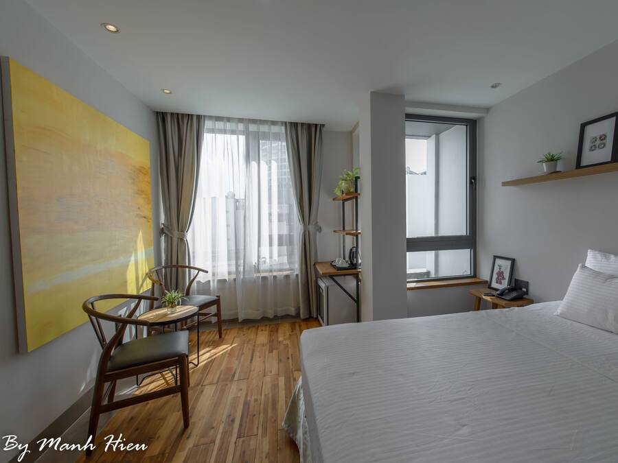 Hotellihuone, jossa on suuri sänky, ruokapöytä tuoleineen, pieni työpöytä tuolilla, ikkuna verhoineen sekä hylly, jossa on valokuvia ja kasvi.
