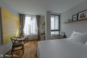 A hotel room with a large bed, a dining table with chairs, a small desk with a chair, a window with curtains, and a shelf with pictures and a plant.