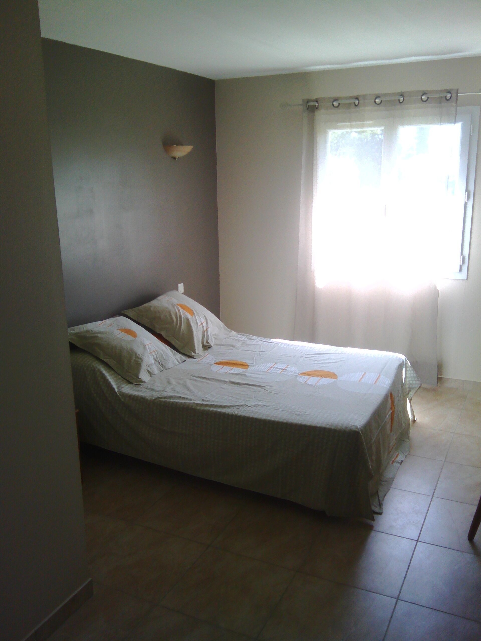 A single bed in a room with a plain wall, a window with curtains, and a tiled floor.