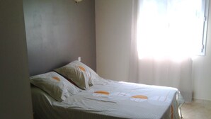 Ein Einzelbett in einem Zimmer mit schlichter Wand, einem Fenster mit Vorhängen und einem Fliesenboden.