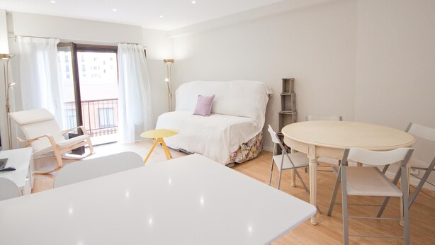 Un dormitorio moderno y bien iluminado con una cama, una pequeña mesa redonda y una silla. Tiene un balcón con vistas a edificios.