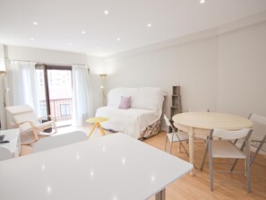 Une chambre moderne et bien éclairée, avec un lit, une petite table ronde et une chaise. Un balcon offre une vue sur les bâtiments environnants.