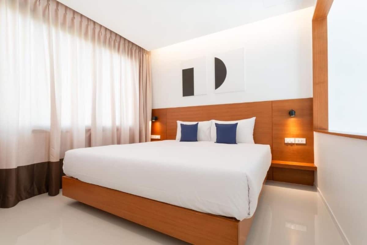 A modern hotel room with a large bed, wooden headboard, two bedside lamps, and a large window with sheer curtains.