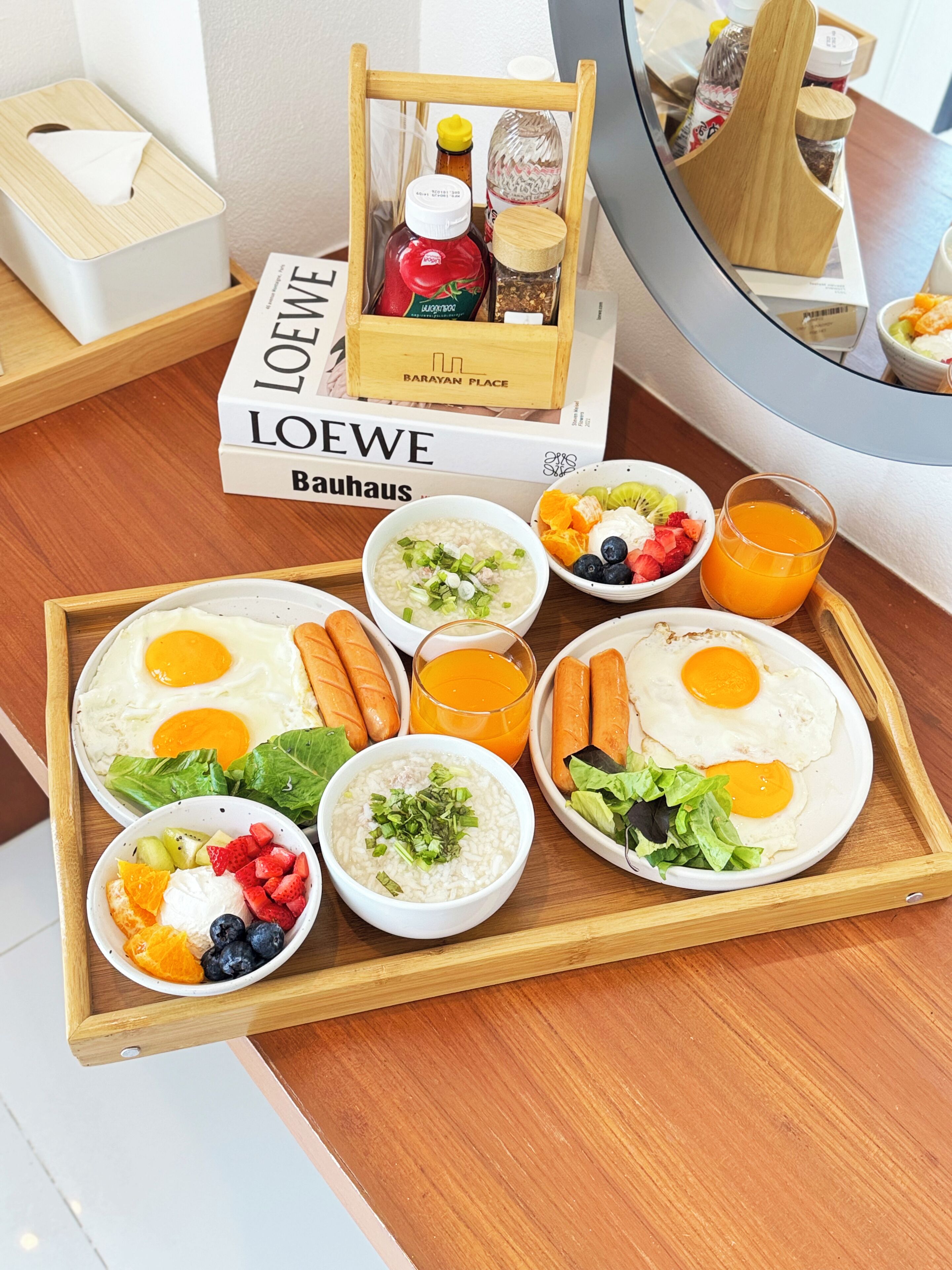 A breakfast tray with eggs, sausages, soup, and fruit on a wooden table.