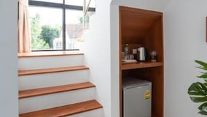 Een trap met houten treden die uitkomt op een plateau, waar een kleine koelkast en een raam met gordijnen staan.
