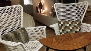 Duas cadeiras de vime branco com almofadas, uma pequena mesa de madeira e uma estante decorativa com velas em um quarto com piso de madeira.