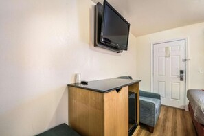 Een kleine kamer met een houten bureau, een aan de muur bevestigde televisie, een deur en een bed met een grijze deken.