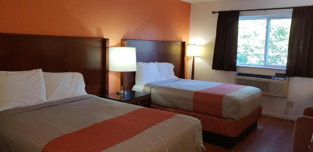 Habitación de hotel con dos camas, una mesita de noche con lámpara, una ventana con cortinas y un equipo de aire acondicionado.
