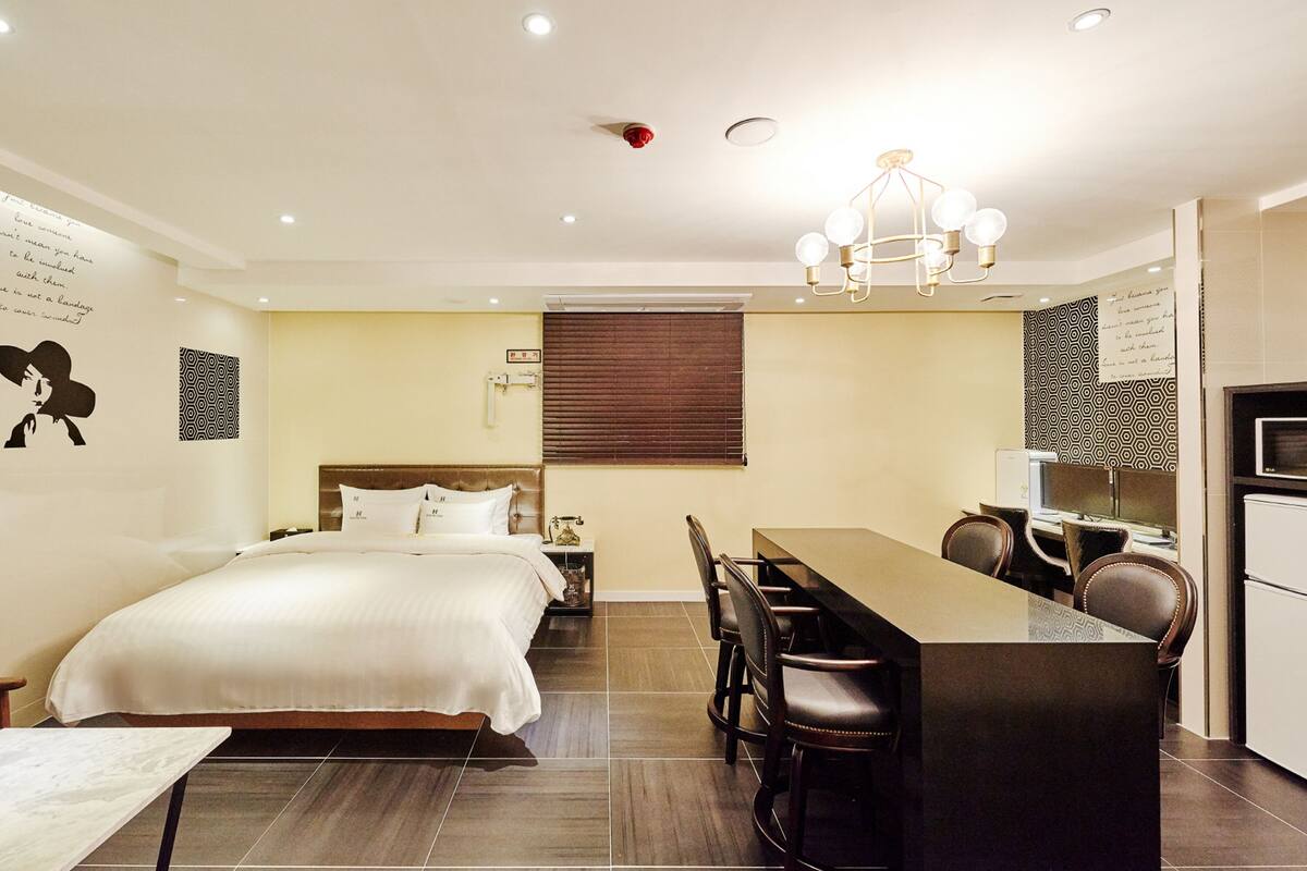 A hotel room with a bed, a dining table with chairs, a small kitchen area, and a ceiling light fixture.