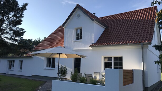 Ett tvåvåningshus med ett rött tegeltak, vita ytterväggar och en liten trädgård med växter och en bänk.