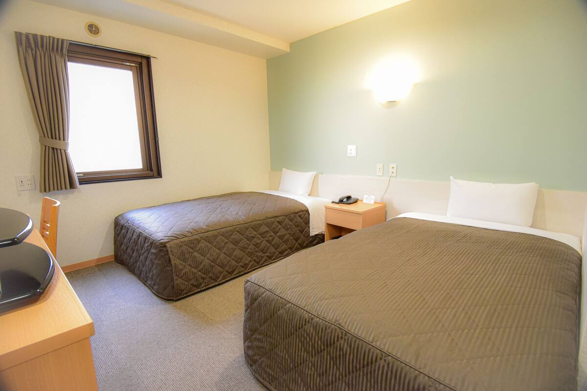 A hotel room with two beds, a desk, a chair, a window with curtains, and a wall-mounted light.