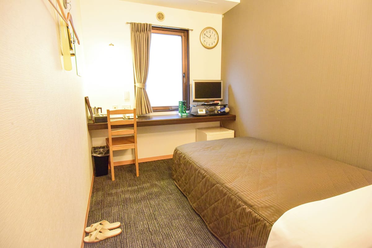 A hotel room with a bed, a desk with a computer, a chair, a window with curtains, and a clock on the wall.