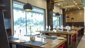 Un restaurant doté de nombreuses tables, chacune mise avec des assiettes, des couverts et des verres. Des lampes suspendues illuminent les tables, et le restaurant possède de grandes fenêtres offrant une vue sur la verdure environnante.