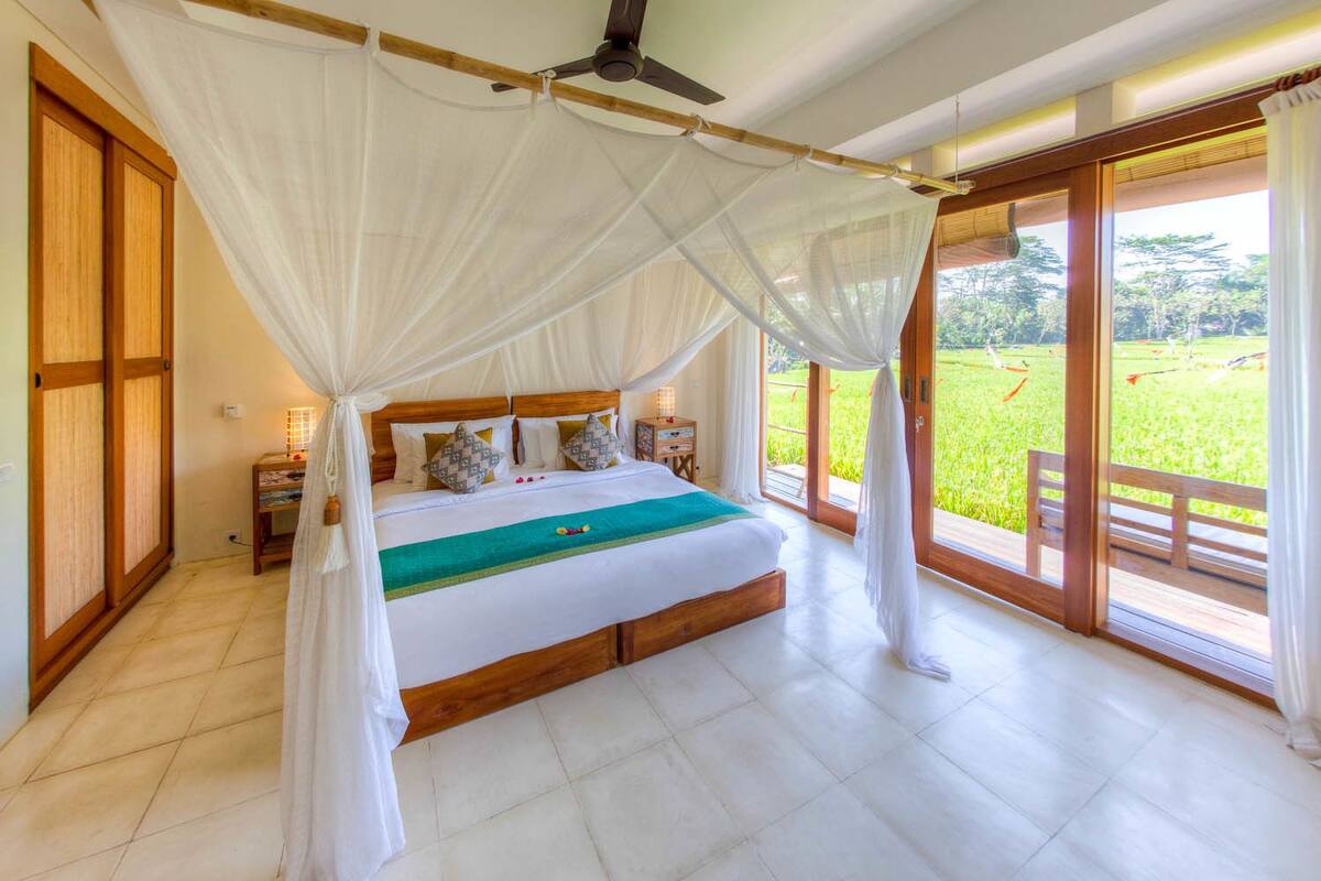 A bedroom with a large bed, wooden headboard, and a canopy. There is a nightstand with a lamp, and a view of a green field through large glass doors.