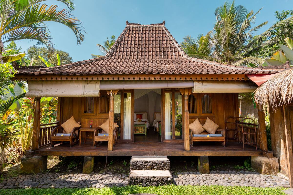 A wooden bungalow with a tiled roof, a porch with seating, and a thatched roof structure.