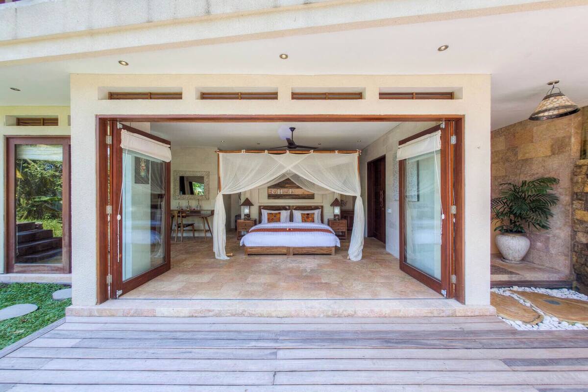 A bedroom with a canopy bed, a desk, and a chair, with a view of the garden through glass doors.