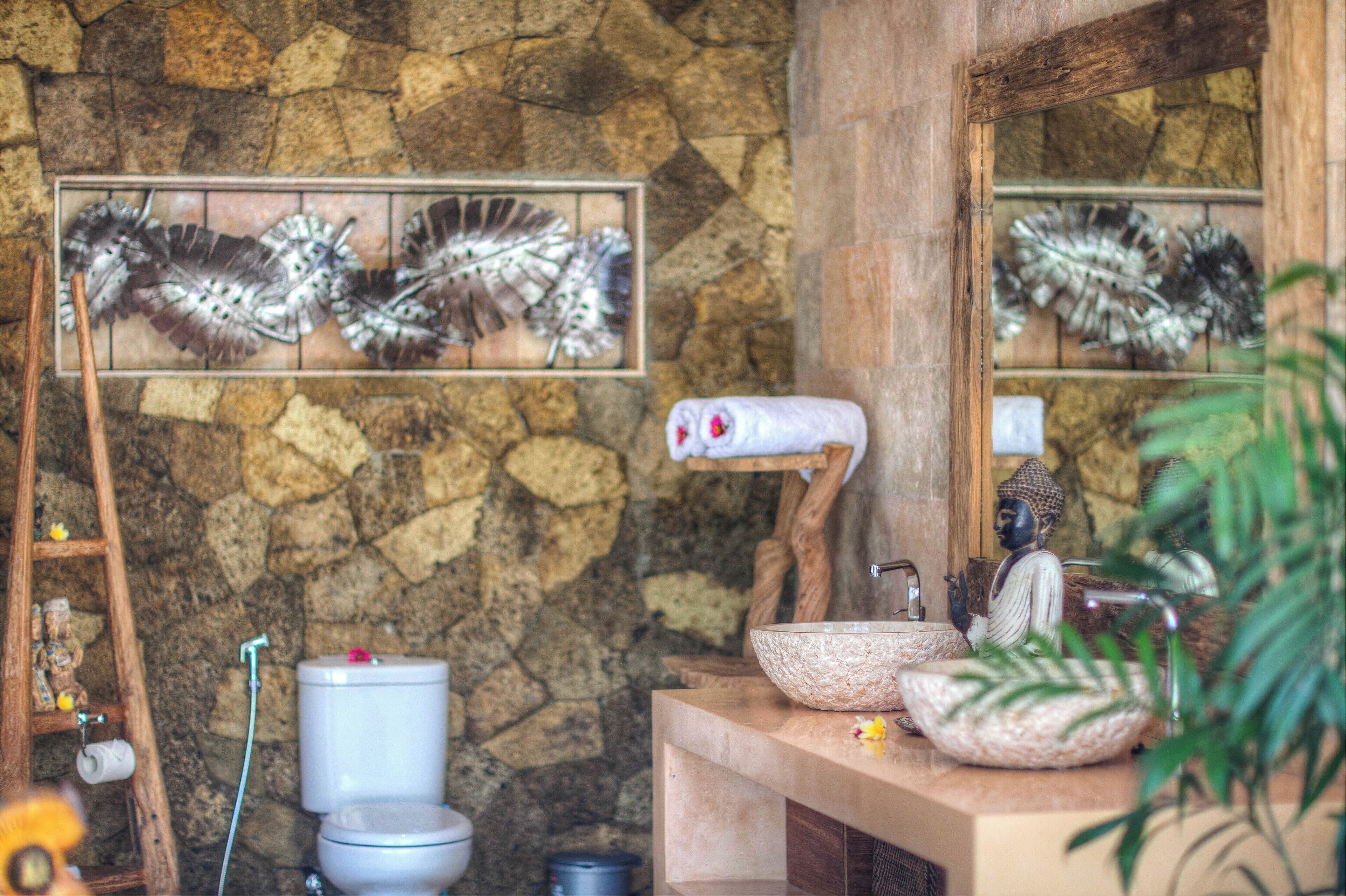 A bathroom with a stone wall, a wooden shelf with a Buddha statue, a large mirror, and a sink with a decorative bowl.