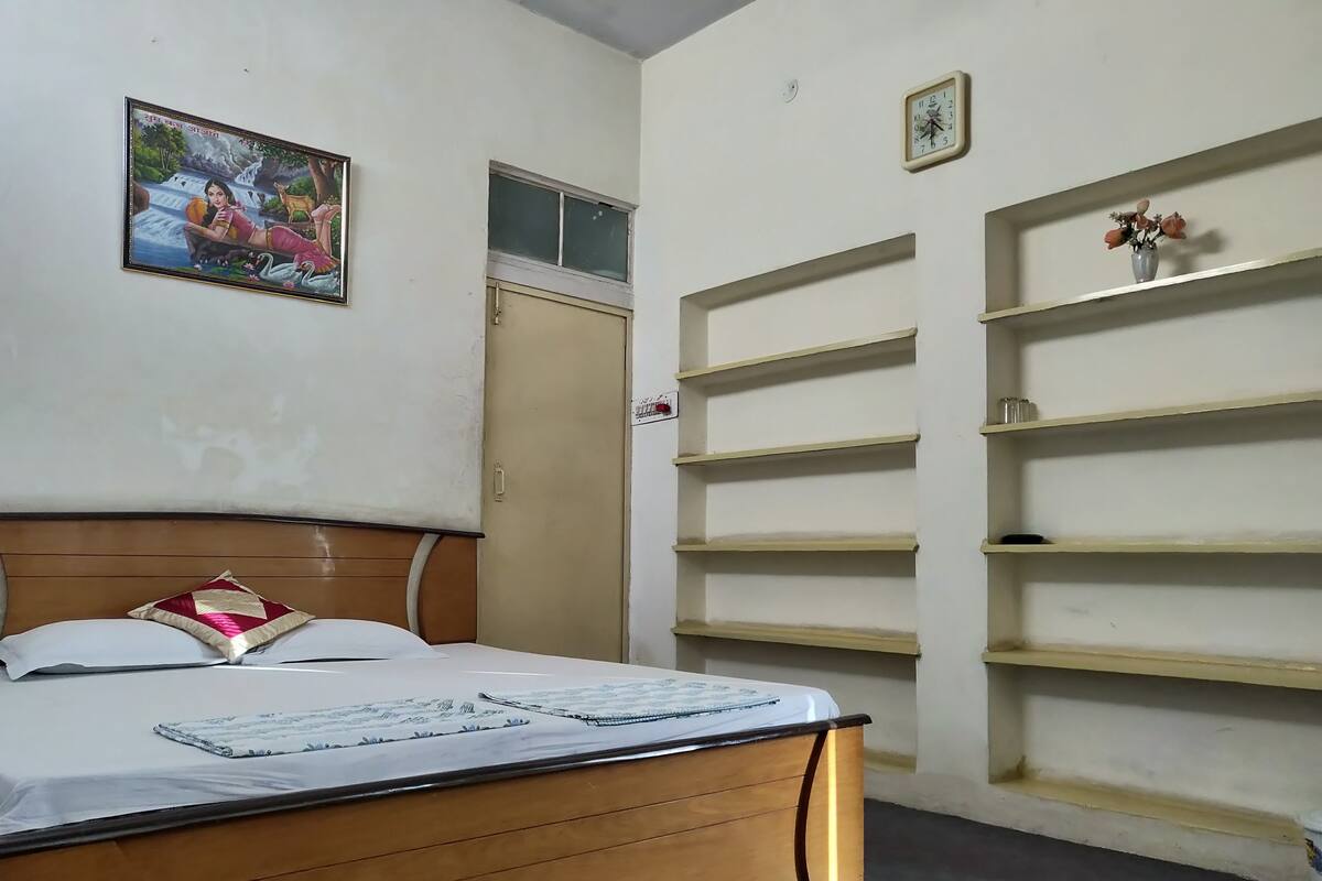 A bedroom with a wooden bed, a wall-mounted shelf with a vase, and a framed picture above the bed.