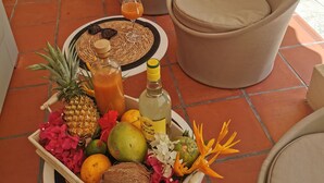 Un área de estar al aire libre con ambiente tropical, con sillas de mimbre y una pequeña mesa con una bandeja de frutas y bebidas.