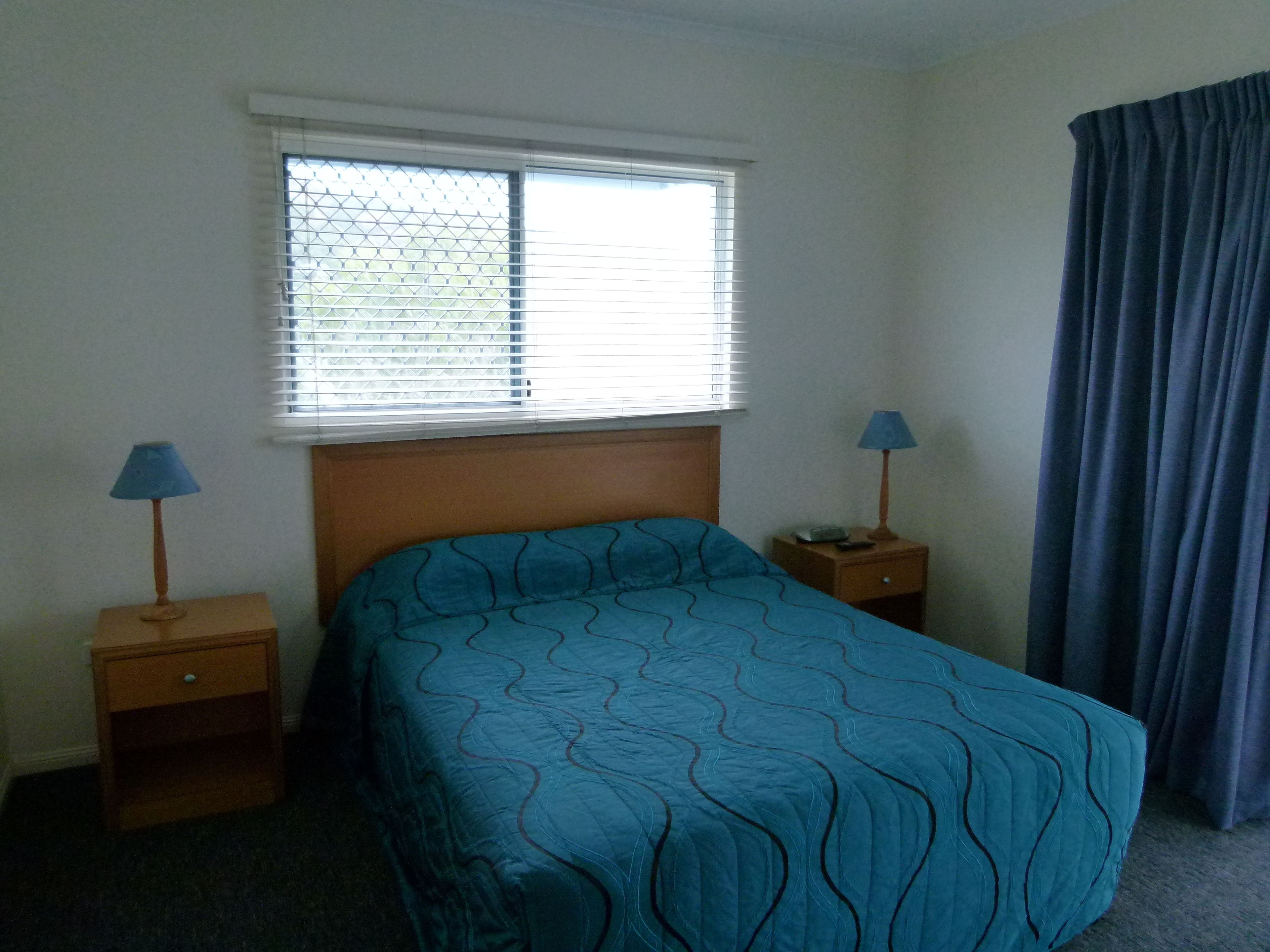 Un dormitorio con una cama individual, dos mesitas de noche, un ventanal con persianas y una cortina.