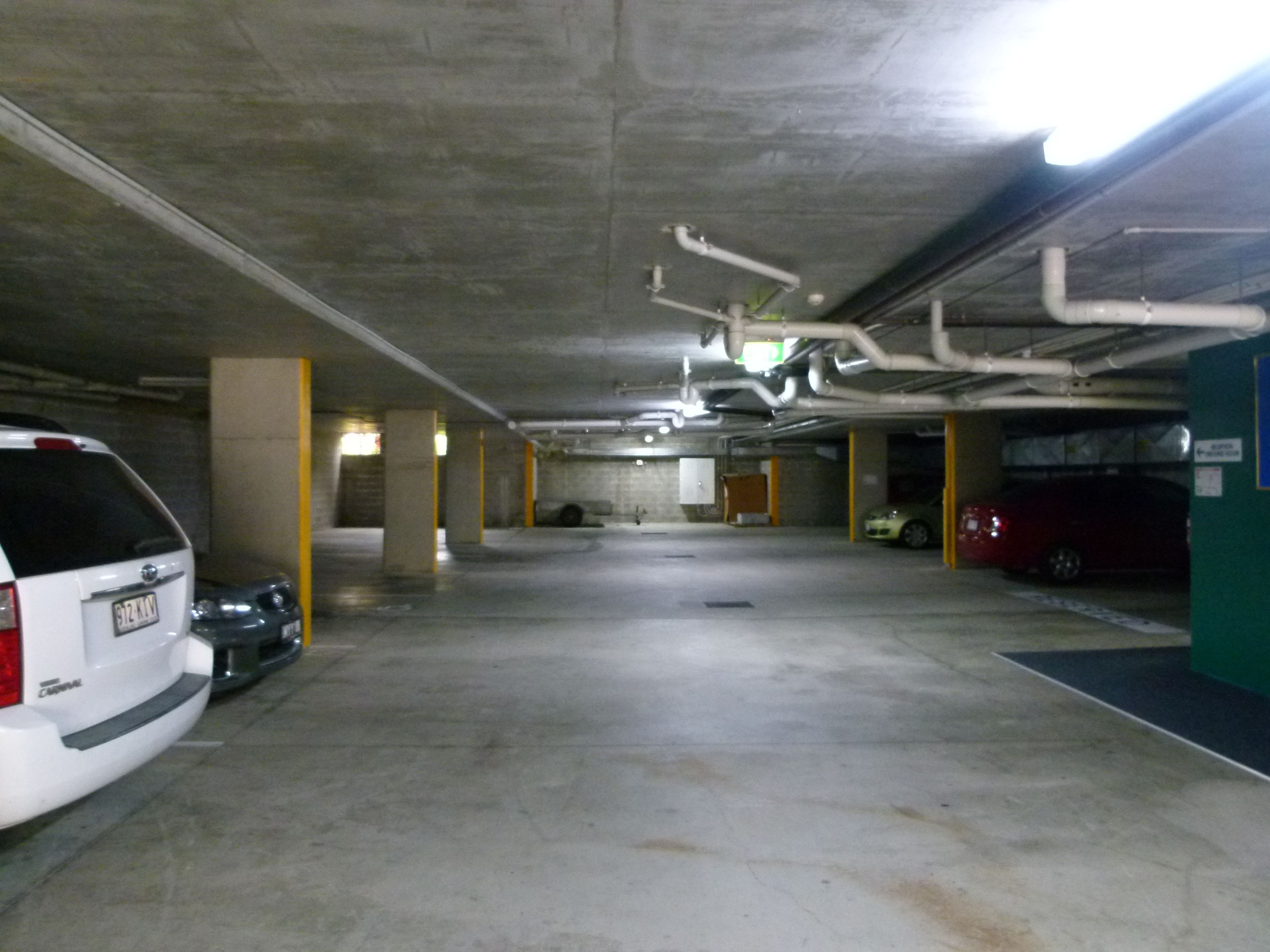 A multi-level parking garage with concrete pillars, overhead pipes, and several parked cars.