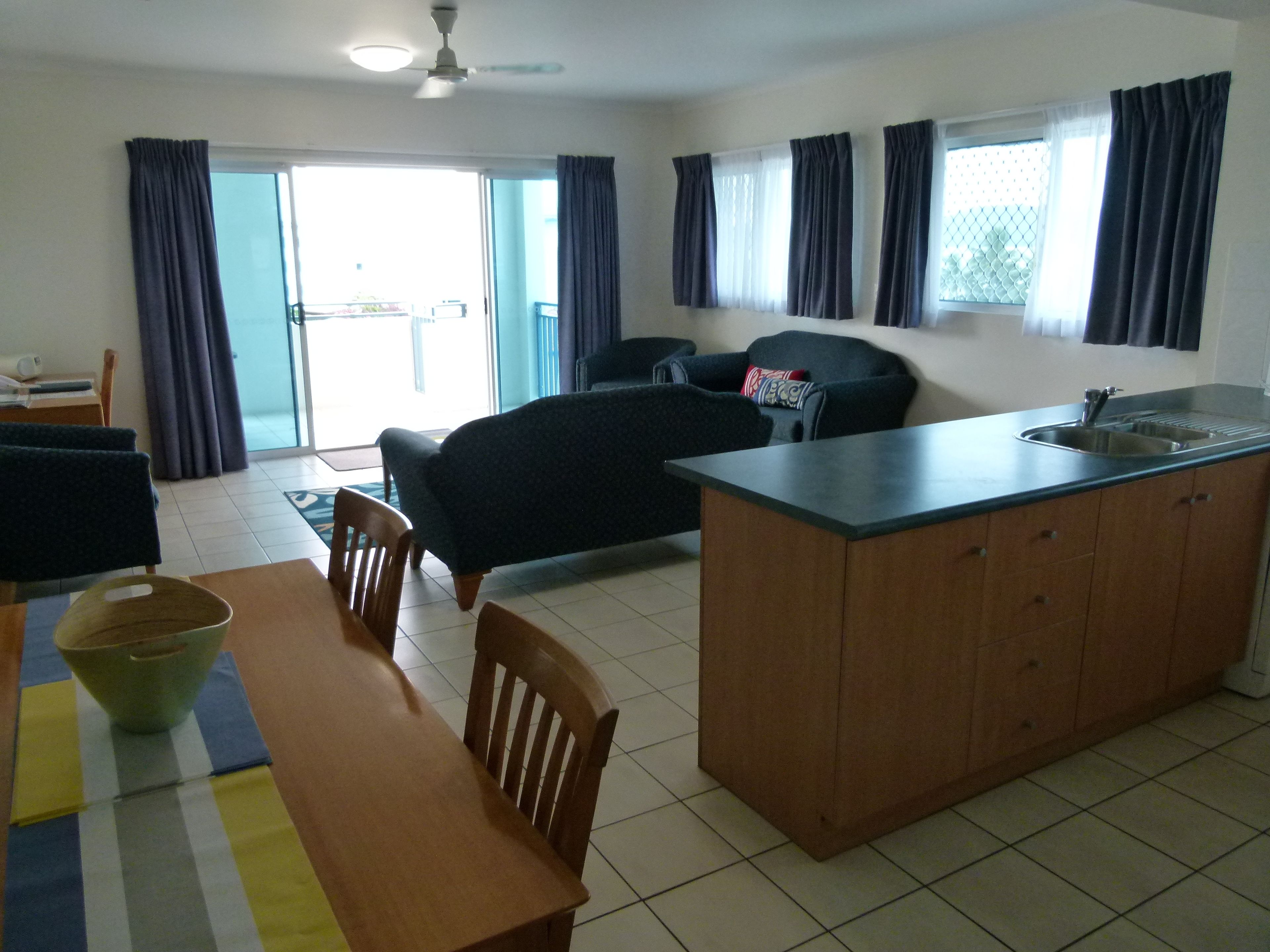 A modern living room with a sofa, a dining table with chairs, a kitchen area with a sink, and a ceiling fan.