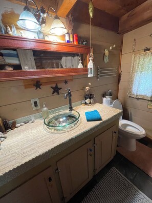 A bathroom with a glass sink, wooden cabinets, a toilet, and a mirror with decorative items. - Rustic Charm close to Portland (Cumberland)