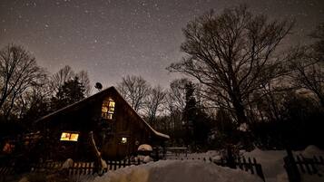 A rustic cabin with a sloped roof, surrounded by snow-covered ground and trees.