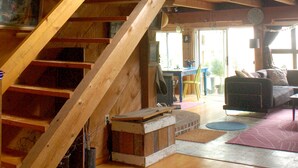 Interior de madera con una escalera que sube a un nivel superior, un comedor y una sala de estar con sofá y cojines.