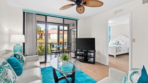 Una sala de estar moderna con una mesa de centro de vidrio, sofás blancos y cojines color turquesa. Se observa un ventilador de techo, un televisor de pantalla plana y un comedor con una mesa y sillas.