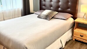 Een netjes opgemaakt bed met een gestuufd hoofdbord, een nachtkastje met een lamp, en een raam met dunne gordijnen.