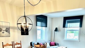 Een eethoek met een houten tafel en stoelen, een bank en een salontafel. Twee ramen met zonwering, een vloerlamp en een kroonluchter.