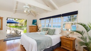 Een slaapkamer met een bed, nachtkastjes, een plafondventilator, uitzicht op de oceaan door grote ramen en een balkon met een stoel en parasol.