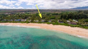Luchtbeeld van een kustgebied met helder turquoise water, een zandstrand en een woonwijk met huizen en groen.