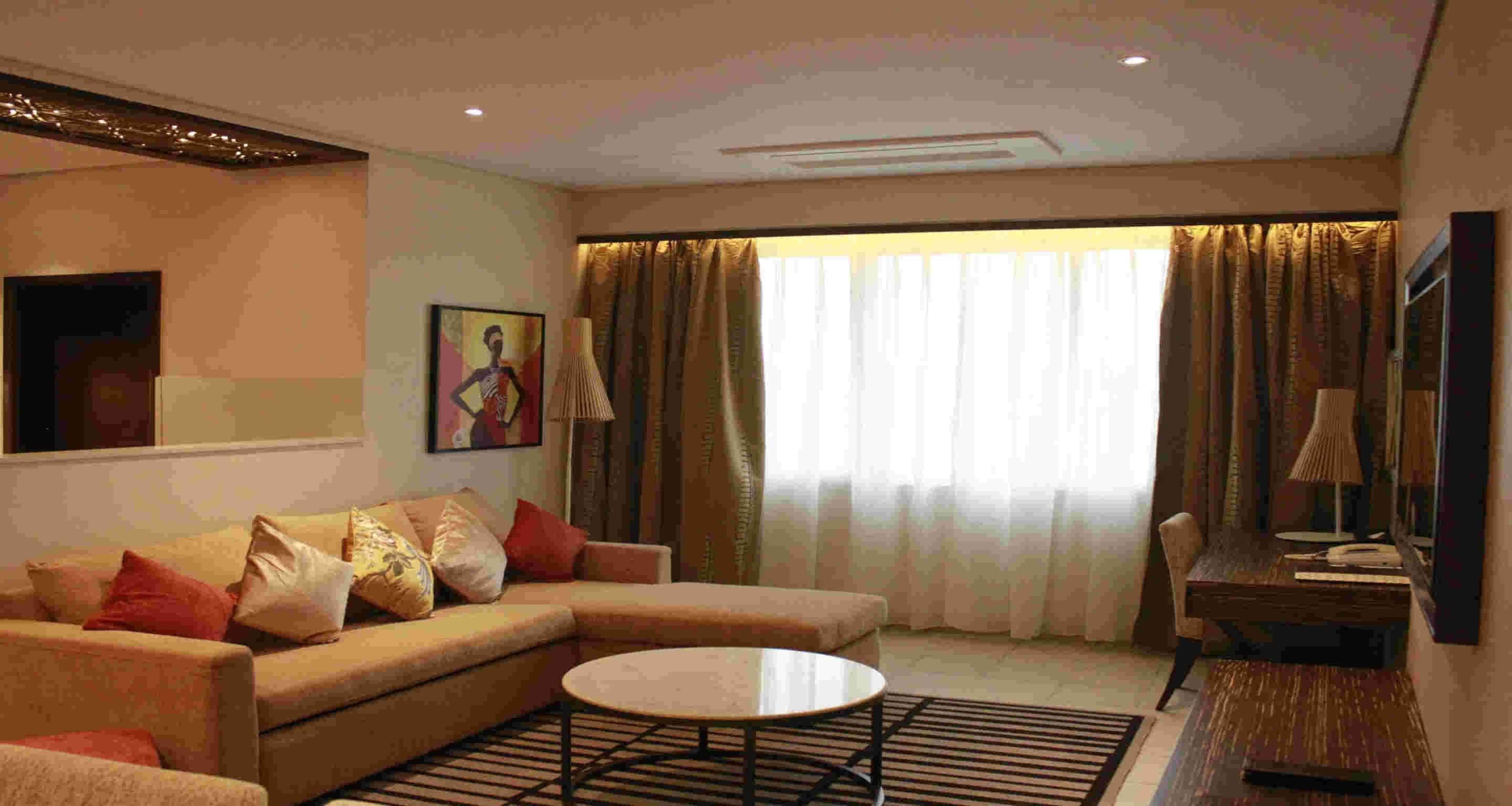 A living room with a sofa, a coffee table, and a lamp. There is a window with curtains and a painting on the wall.