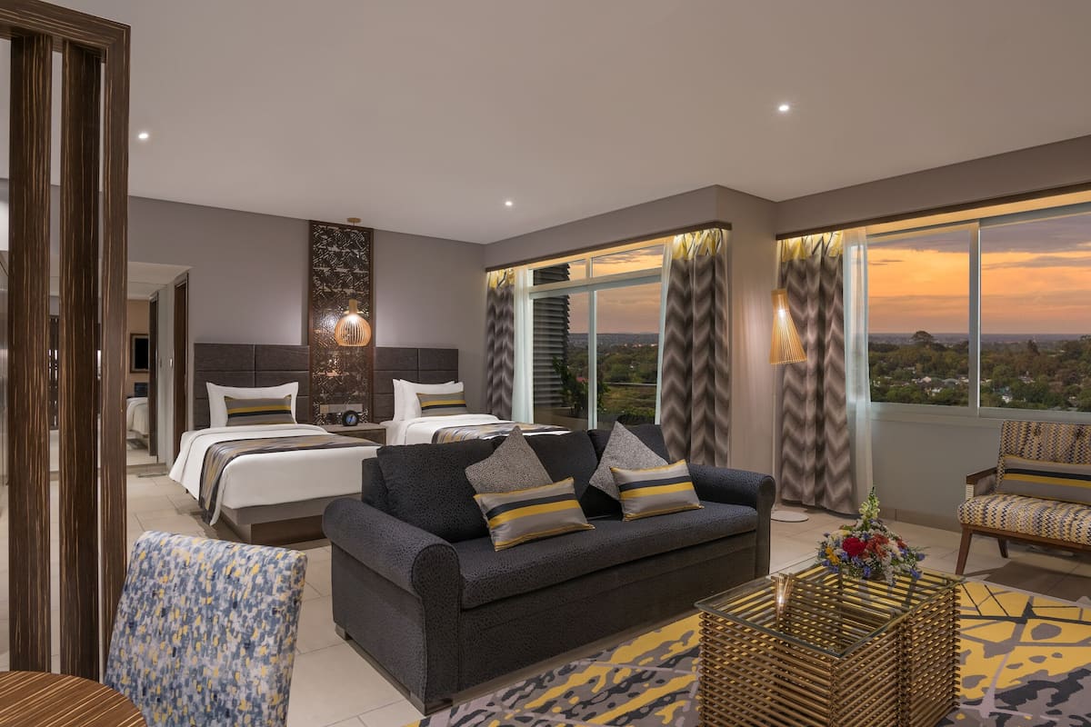 A modern hotel room with a large bed, a sofa, and a view of the cityscape at sunset.