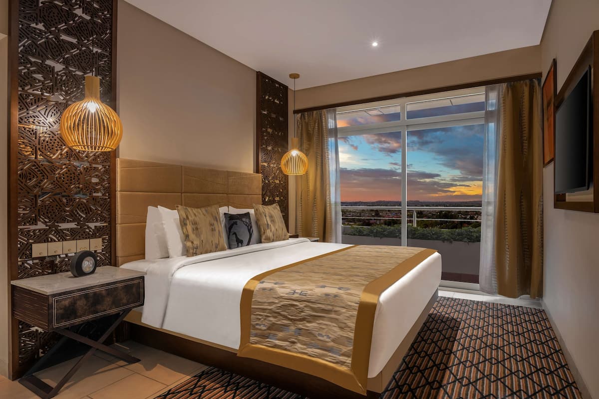 A modern hotel room with a large bed, a decorative wall panel, a nightstand with a clock, and a large window with a sunset view.