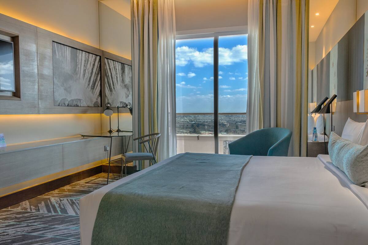 A hotel room with a large bed, a desk with a chair, a view of the city, and a window with curtains.