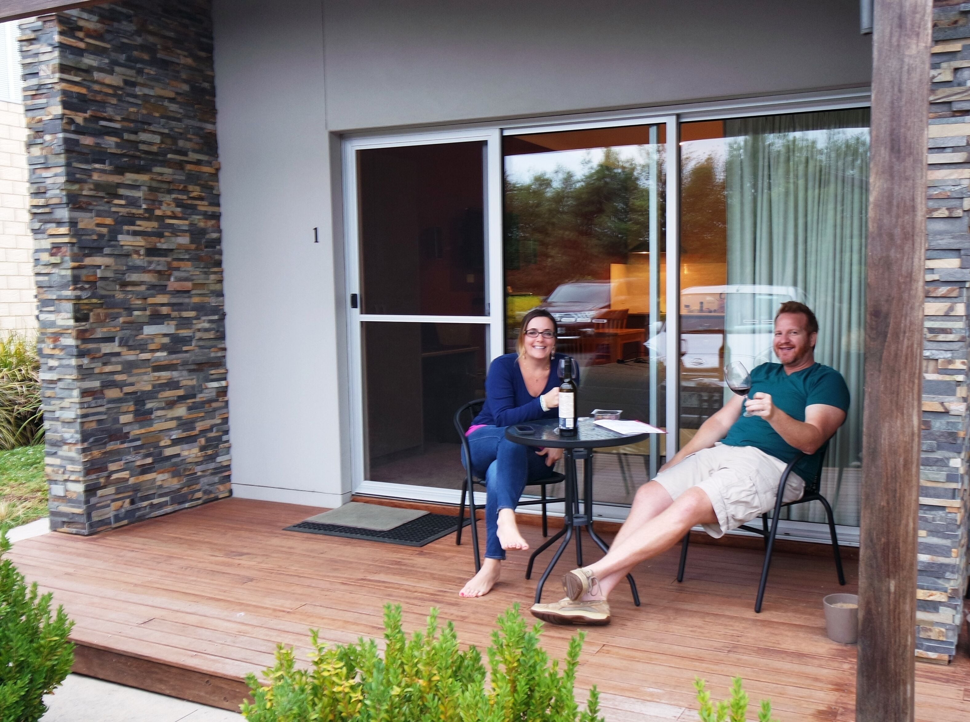 Un patio moderno al aire libre con un muro de piedra, un ventanal de vidrio que da a un espacio cubierto y una terraza de madera.