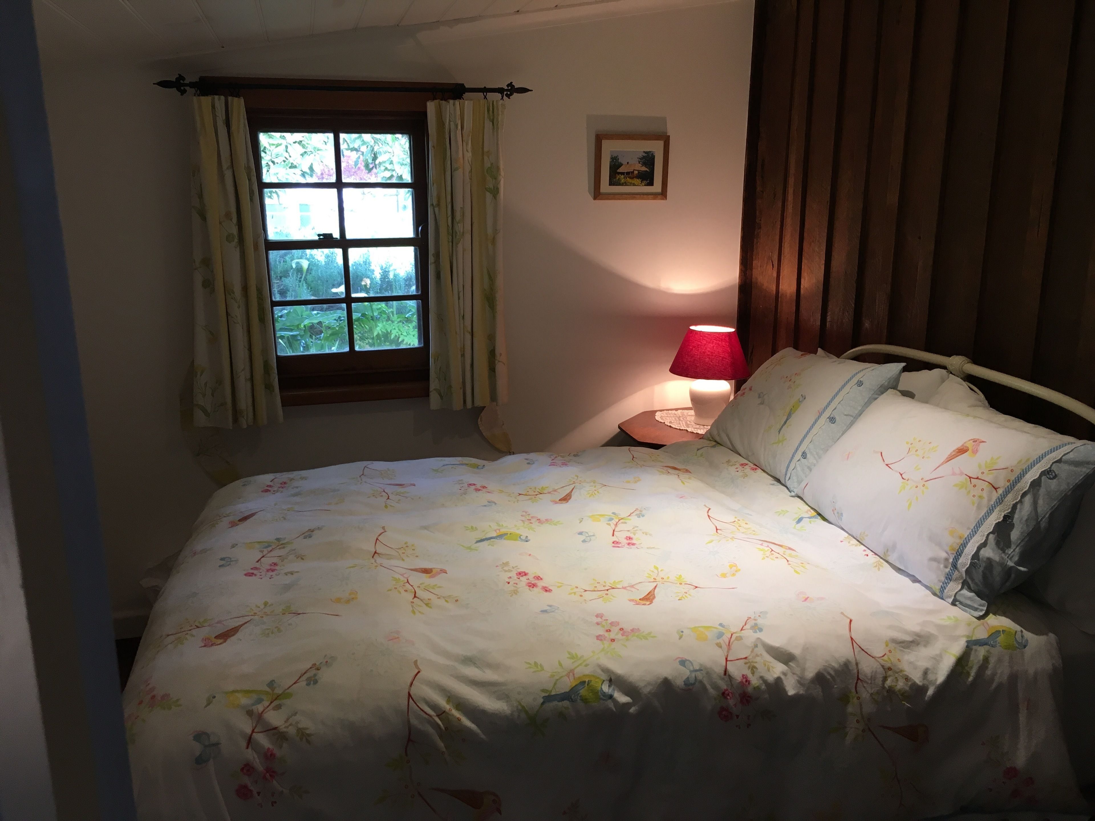 A bedroom with a bed, a window with curtains, a bedside lamp, and a small picture on the wall.