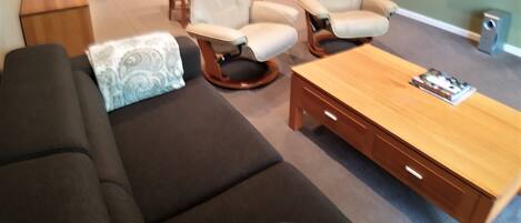 A modern living room with a dark sofa, two reclining chairs, a wooden coffee table, and a kitchen in the background.