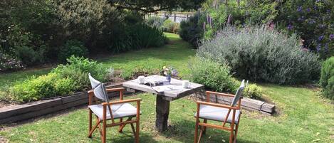 Un área de estar al aire libre con dos sillas de madera y una mesa, rodeada de follaje exuberante y plantas florecientes.