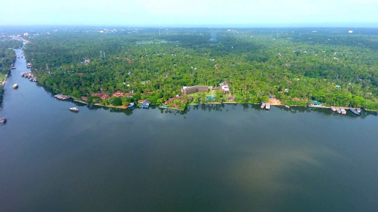 An aerial view of a river with boats, dense greenery, and a large building with multiple structures.
