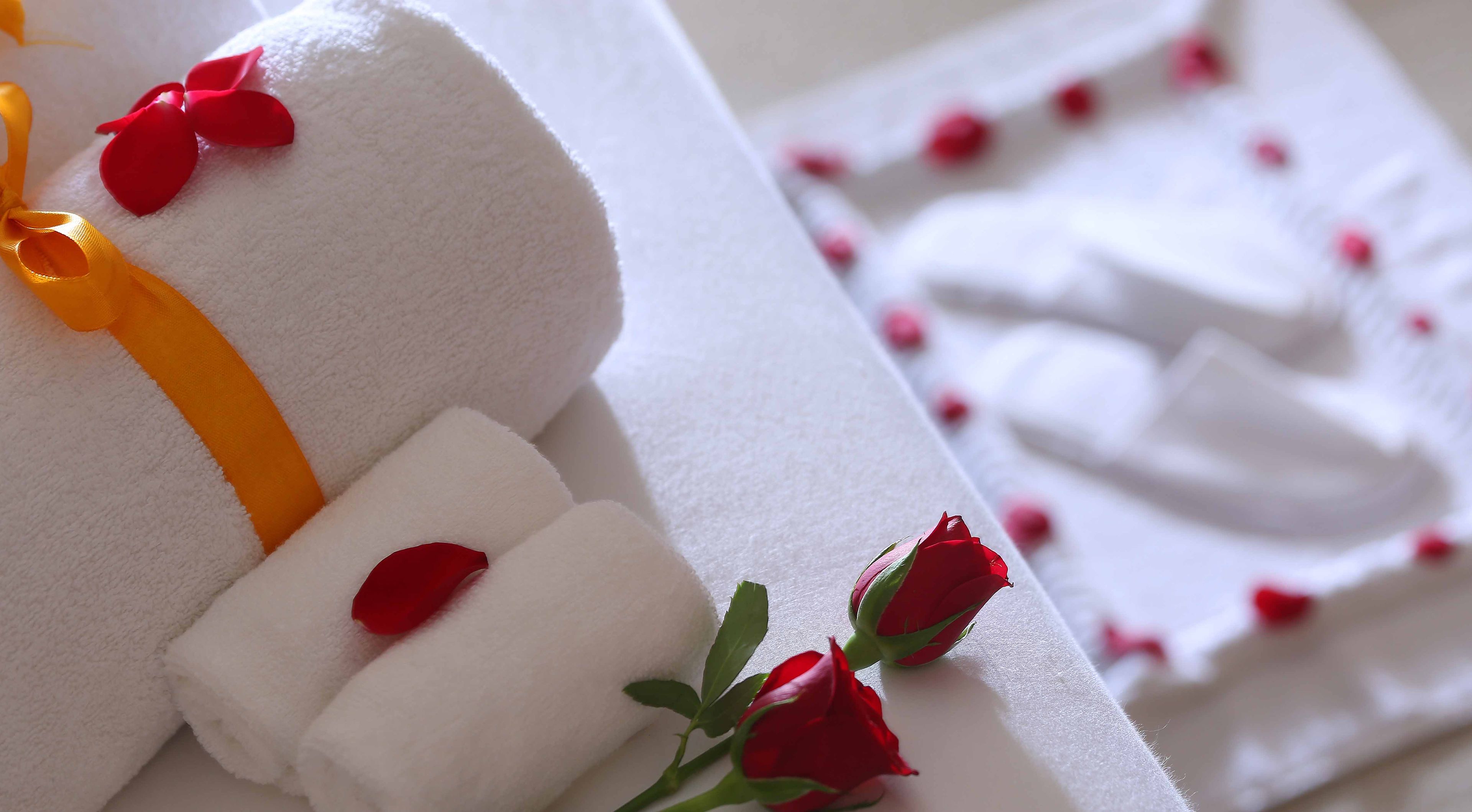 A neatly folded white towel with a yellow ribbon and red rose petals.
