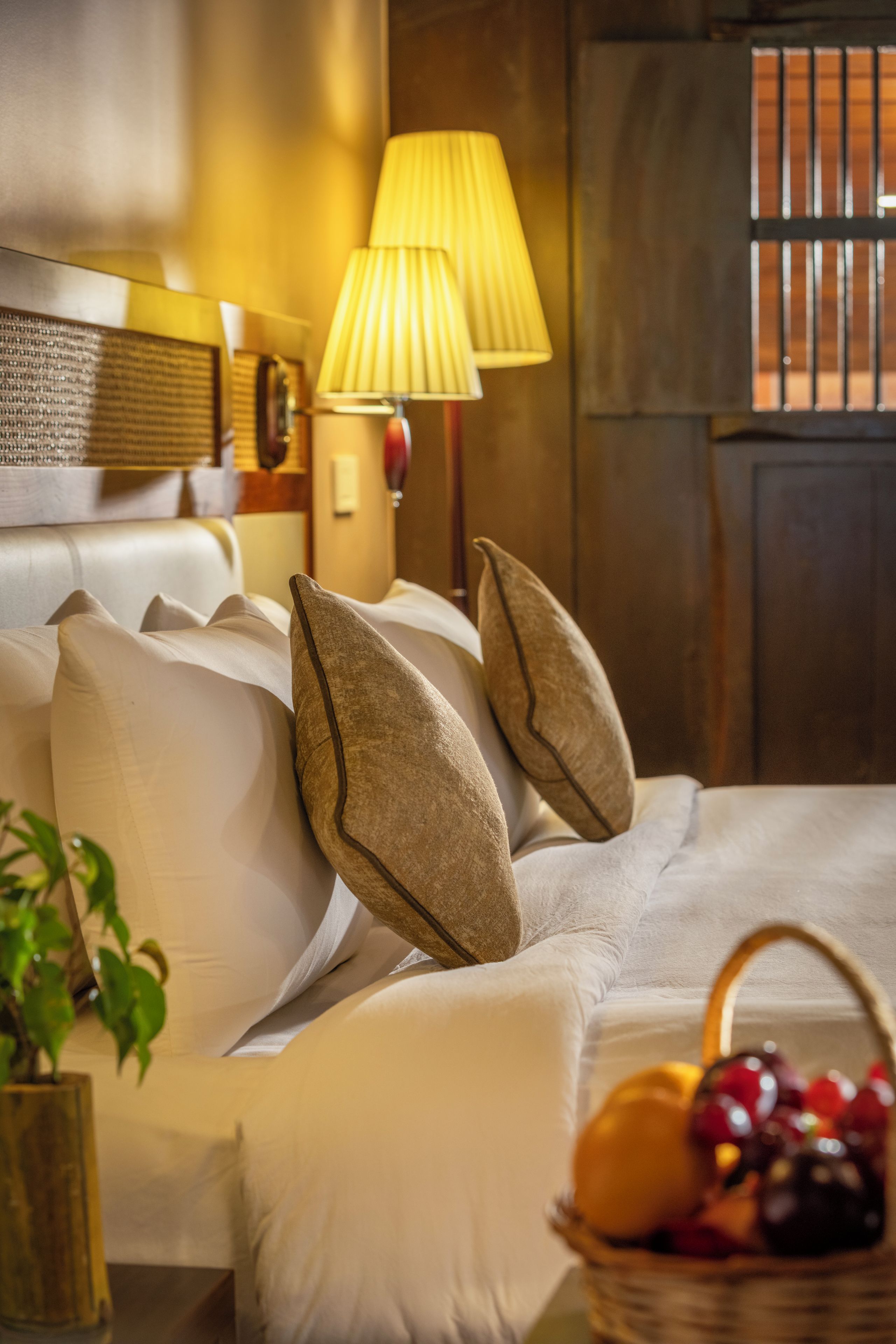 A hotel room with a bed, pillows, a lamp, a fruit basket, and a window with bars.