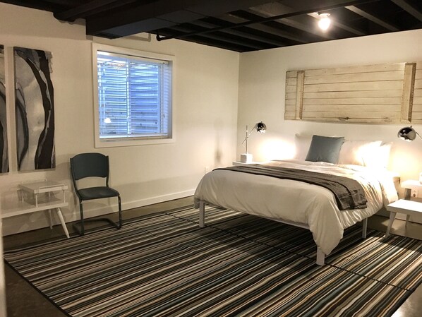 A bedroom with a bed, a chair, a window with blinds, a lamp, and a wall-mounted shelf.