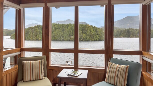 A cozy cabin interior with wooden beams, two upholstered chairs with striped pillows, a small table, and large windows offering a view of a lake and forest. - Waterfront - Black Rock Landing (Ucluelet)