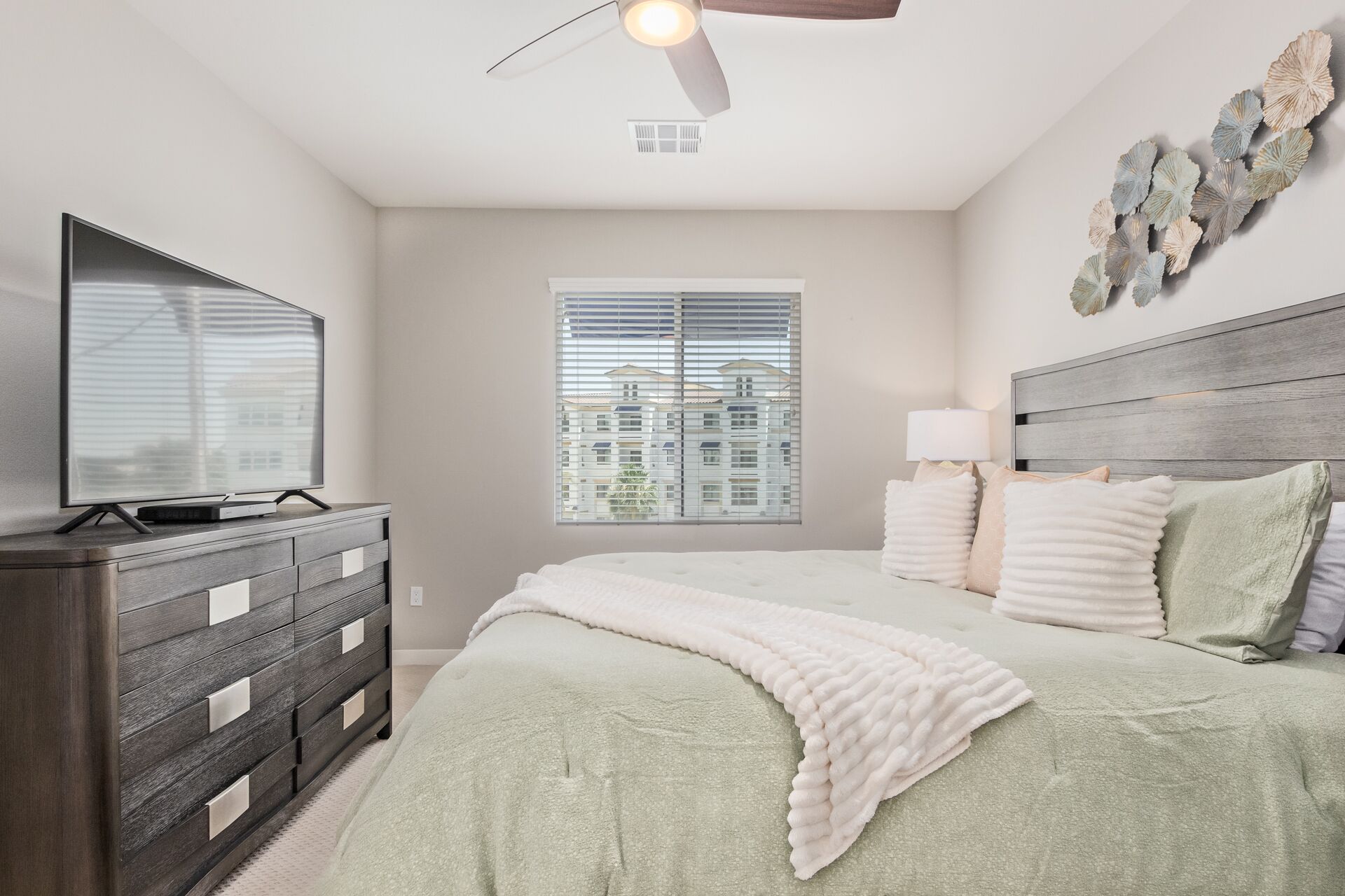 A bedroom with a bed, a dresser, a ceiling fan, a window with blinds, and a view of buildings outside.